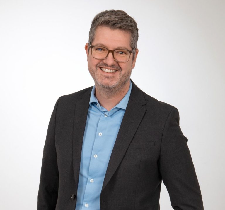 Mann in Anzug mit Brille, lächelnd vor neutralem Hintergrund.